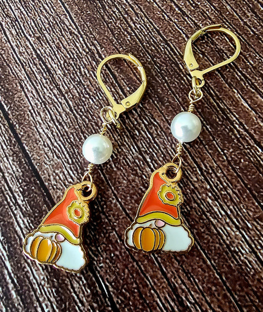 A pair of gold leverback earrings featuring white pearl beads and small gnome charms with red, orange, and white enamel details, displayed on a dark wood background.